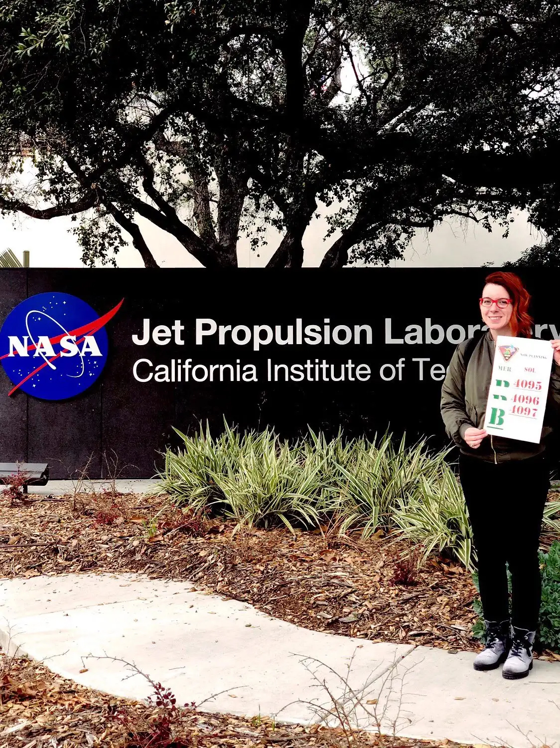 Tanya Harrison standing at the entrance to the NASA Jet Propulsion Laboratory holding a sol sheet from her time on the Opportunity rover mission